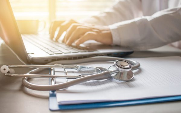 Doctor working on a laptop with a stethoscope on the desk next to him.