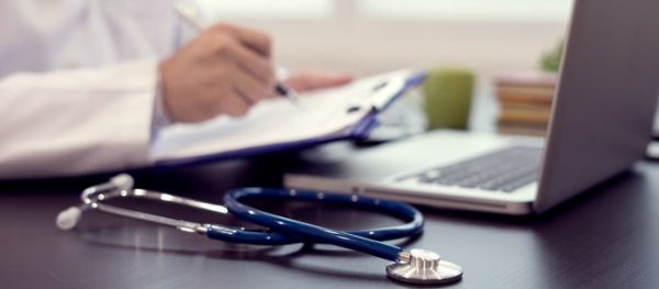 A stethoscope sits on a desk beside a doctor writing on papers on a clipboard in front of a laptop.