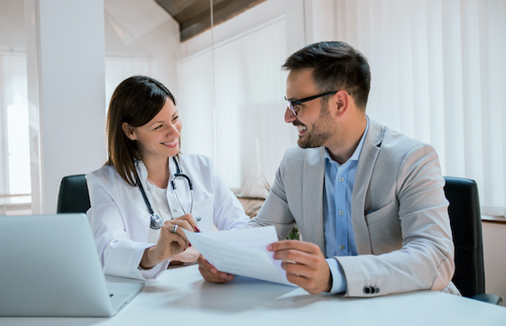 A doctor sitting at a desk in front of a laptop smiling at a man holding papers.