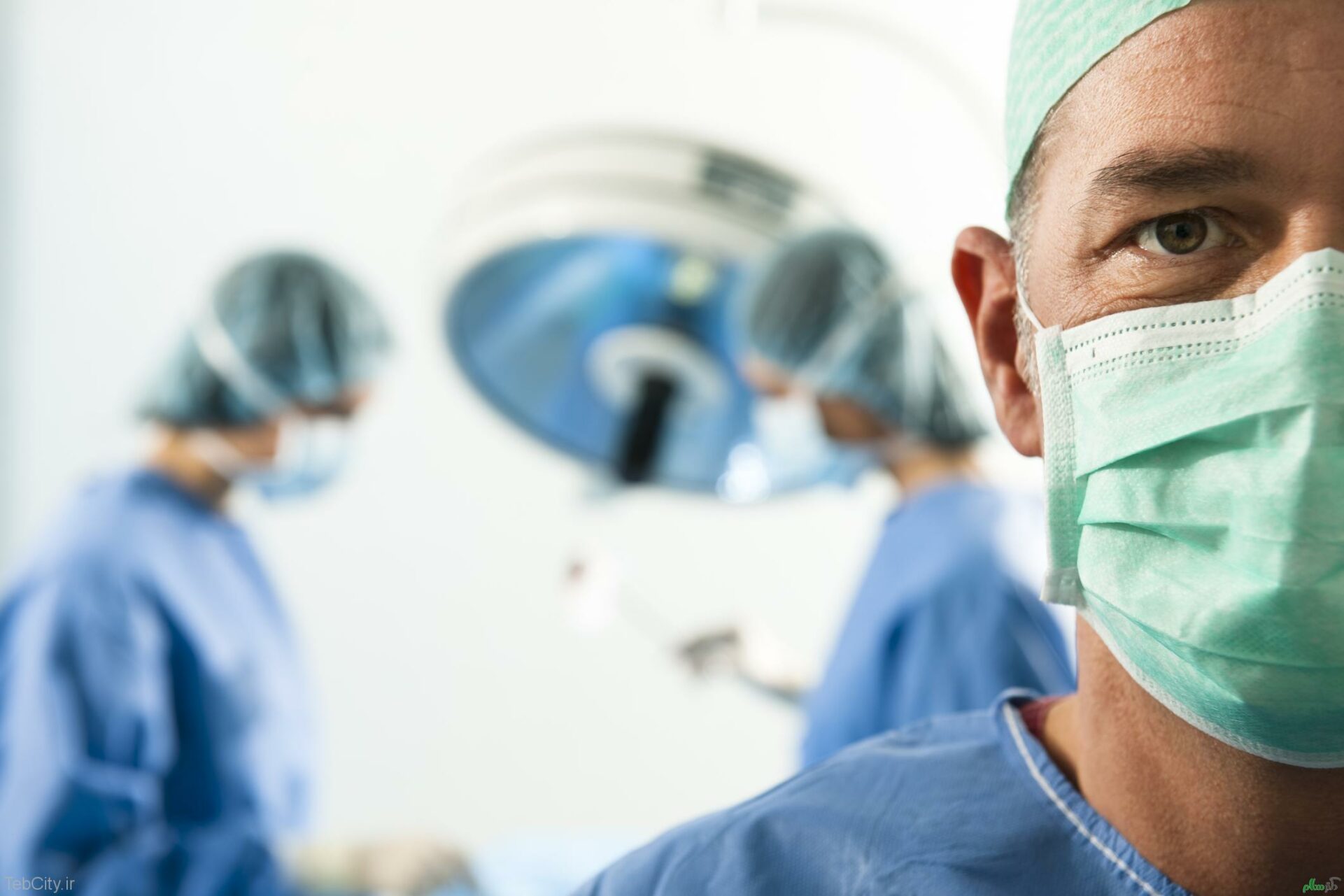 surgeon looking into a camera with a mask on while other surgeons are behind him working.