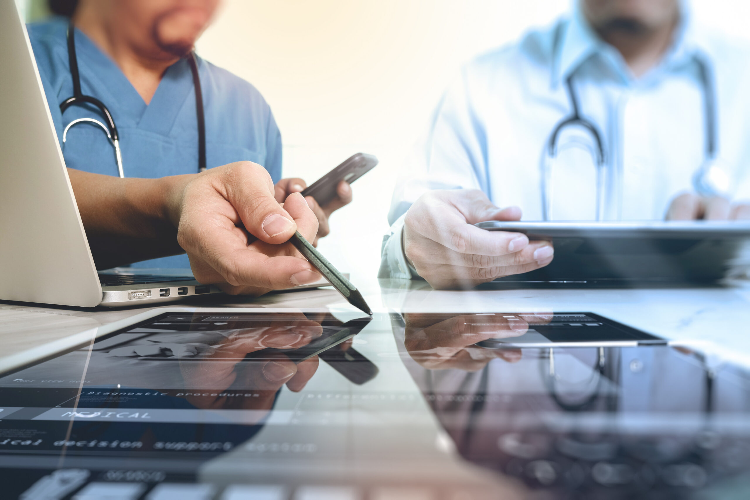 Two doctors at a desk looking over tablets and a laptop.