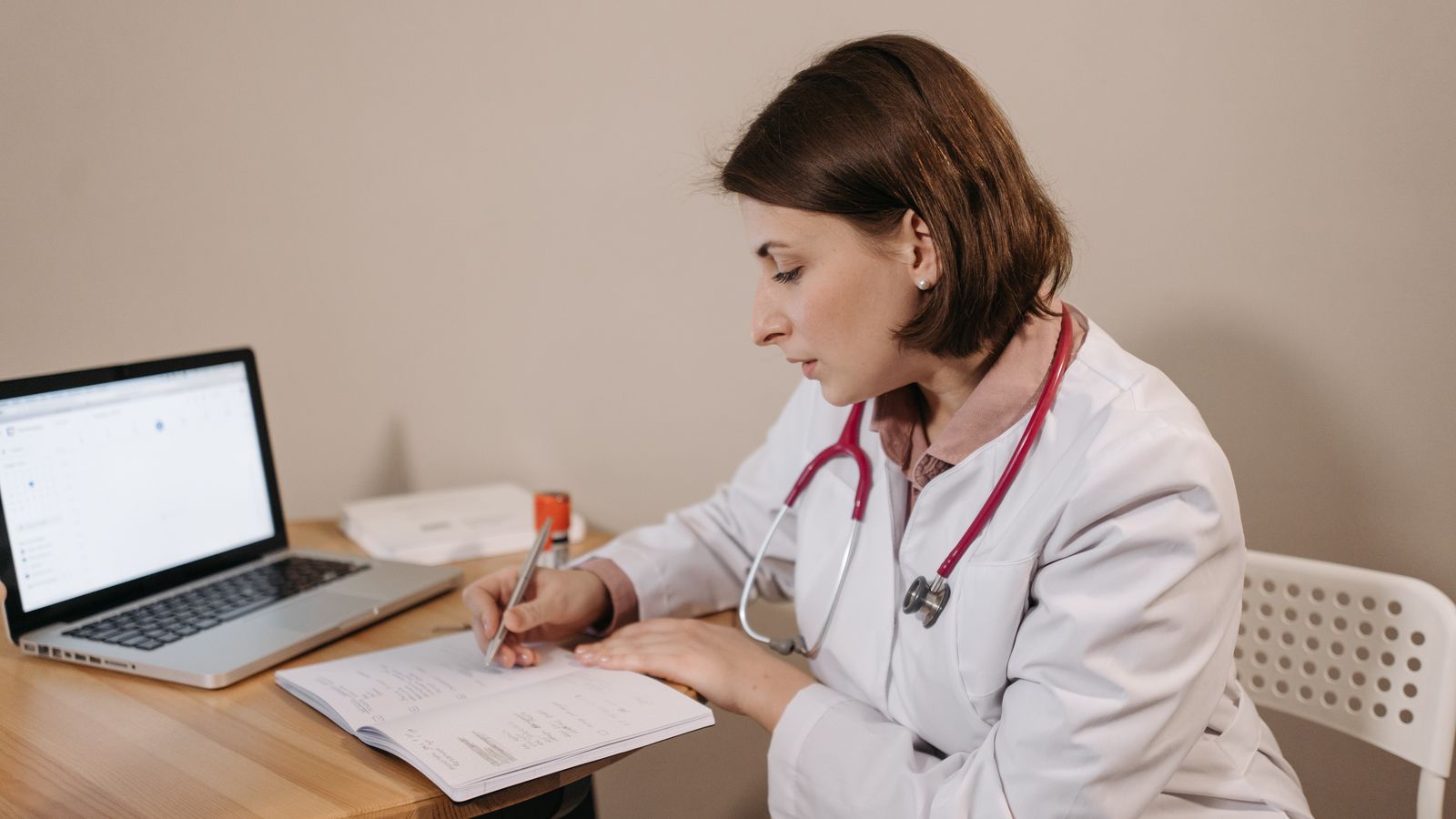 A physician in a lab coat reviewing documents at a laptop, representing provider credentialing paperwork