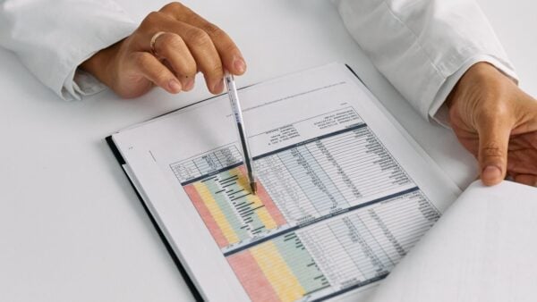 Hands in a white coat pointing a pen at a printed table of clinical data, representing a medical practice analyzing its exposure to the 2027 Medicare physician fee schedule